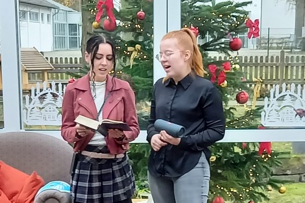 Zwei junge Frauen stehen singend vor einem geschmückten Weihnachtsbaum; eine hält ein Gesangbuch, die andere eine Musikbox.