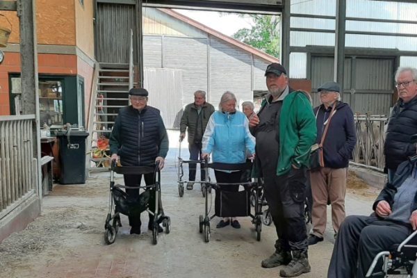 Eine Seniorengruppe der Tagespflege mit Rollatoren und einem Rollstuhl steht gemeinsam mit dem Landwirt Karsten von der Ahe im modernen Kuhstall eines landwirtschaftlichen Betriebs.